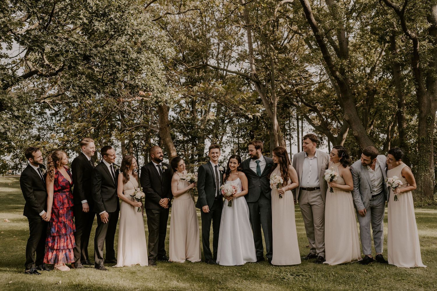 Wedding party poses together outdoors under tall trees in formal attire.