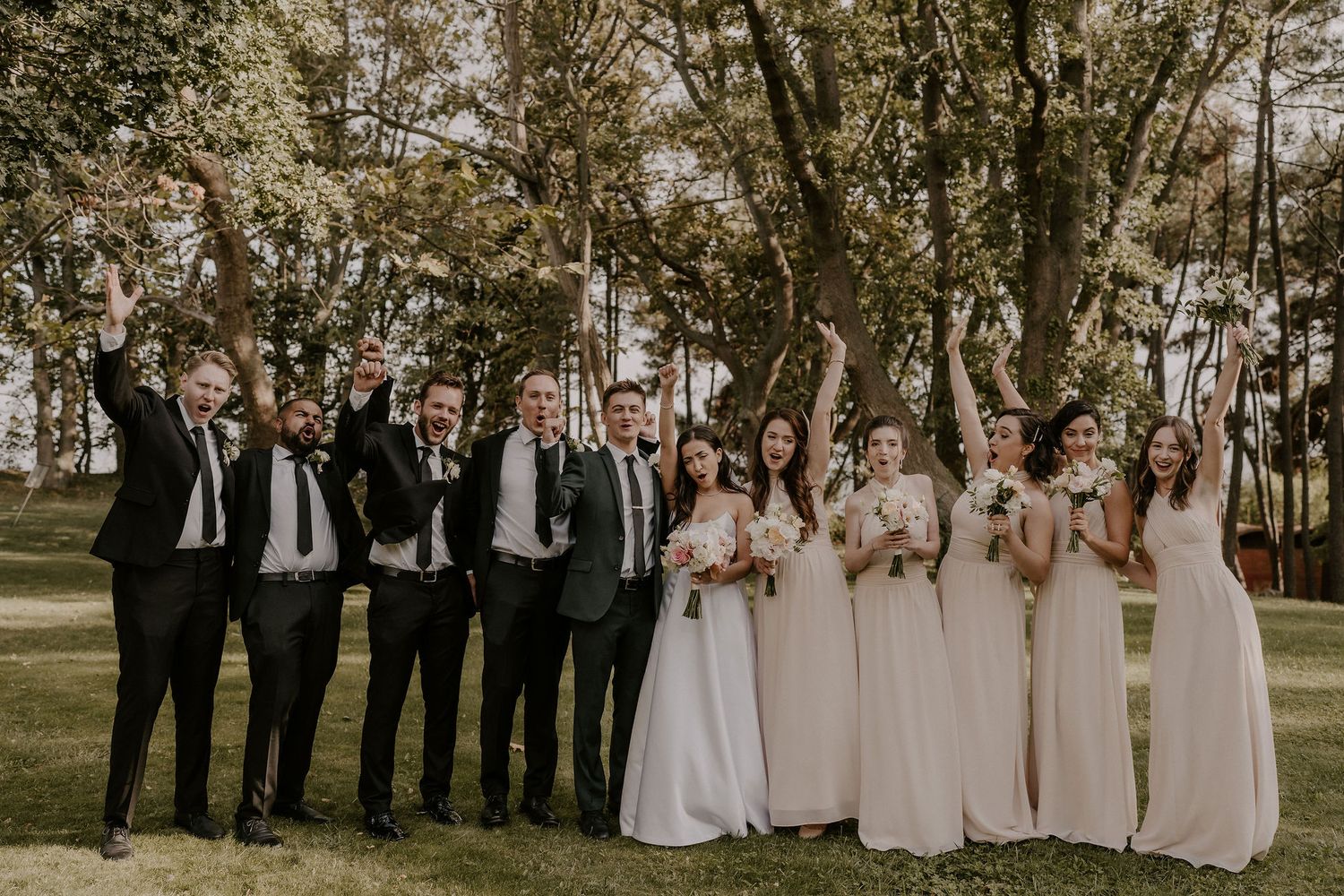 Groomsmen in black suits and bridesmaids in light dresses pose together under trees.