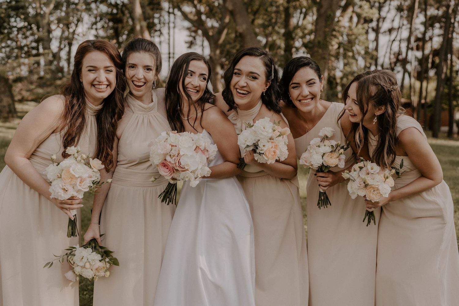 Bridesmaids in blush pink dresses pose together holding white and pink bouquets at an outdoor wedding.
