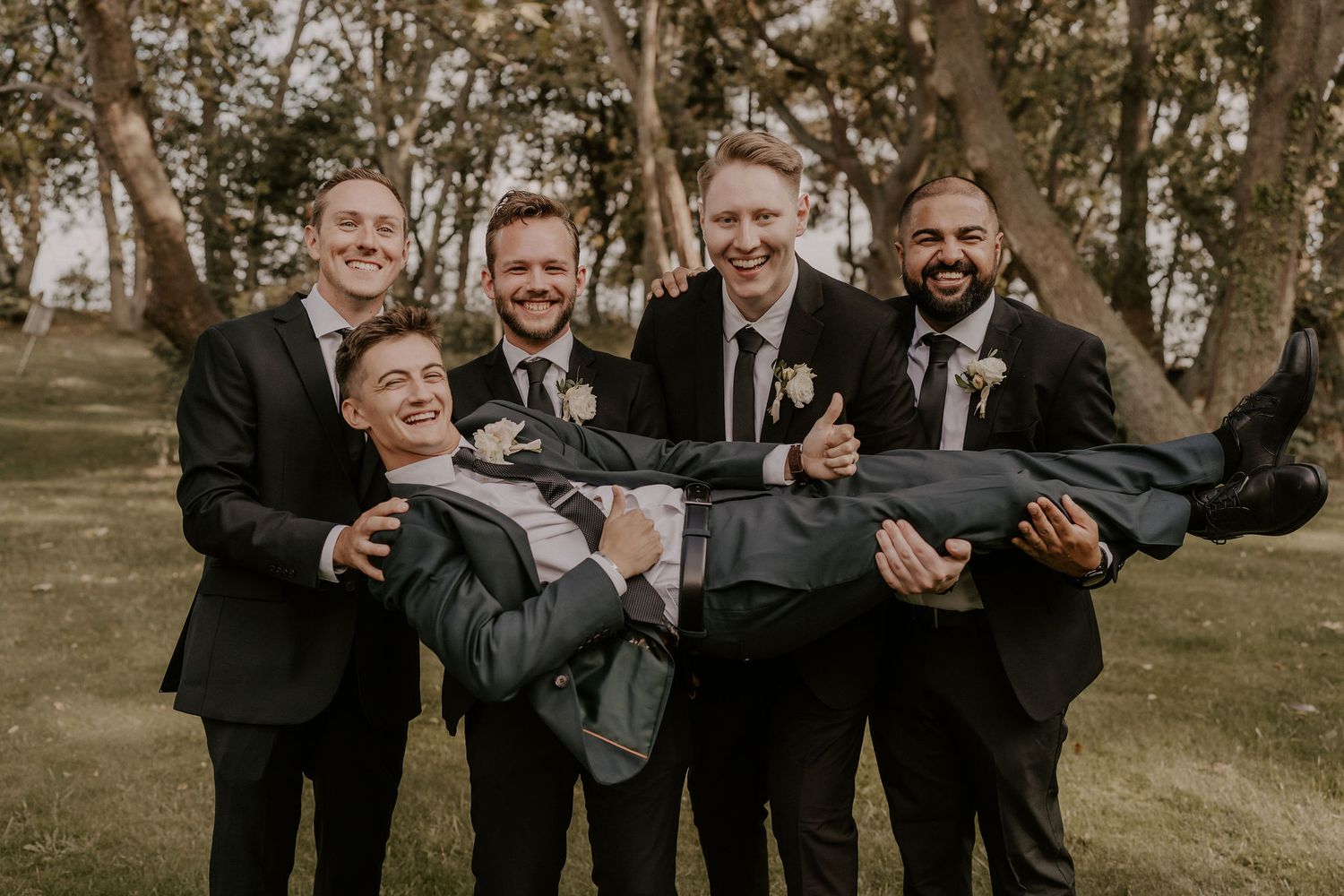 Groomsmen in dark suits share a playful moment while posing outdoors.