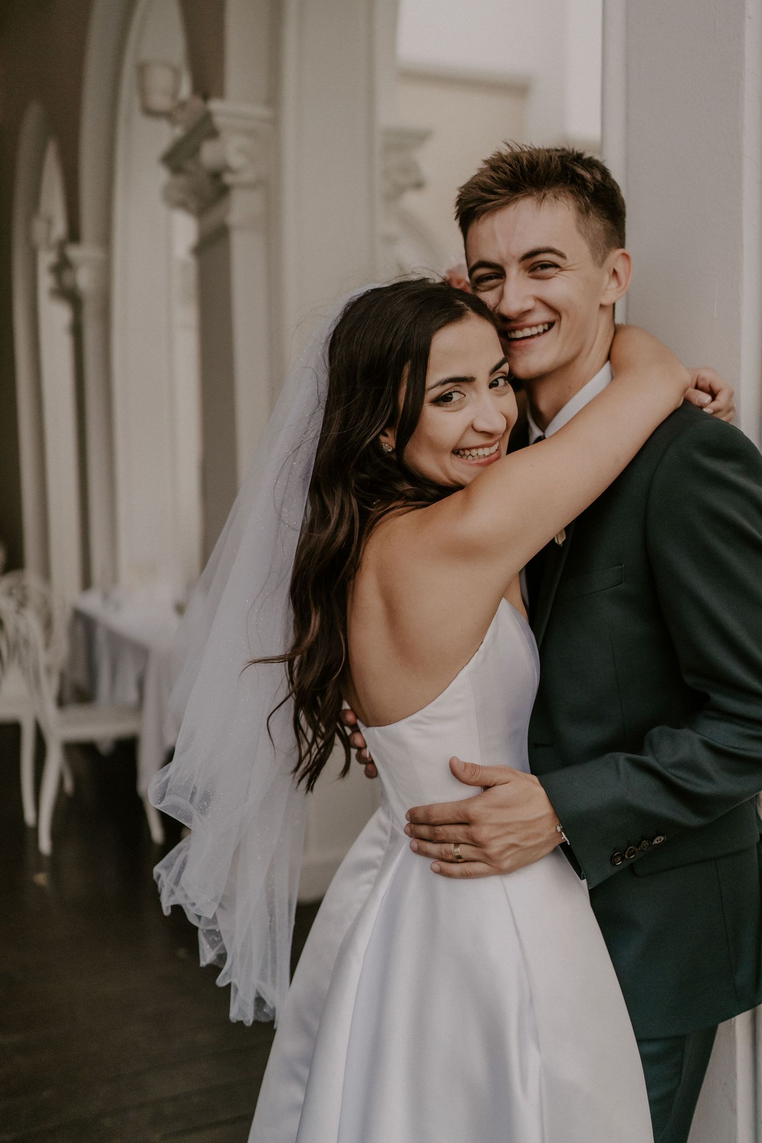 A couple shares joyful moments together under white columns during their wedding photoshoot.