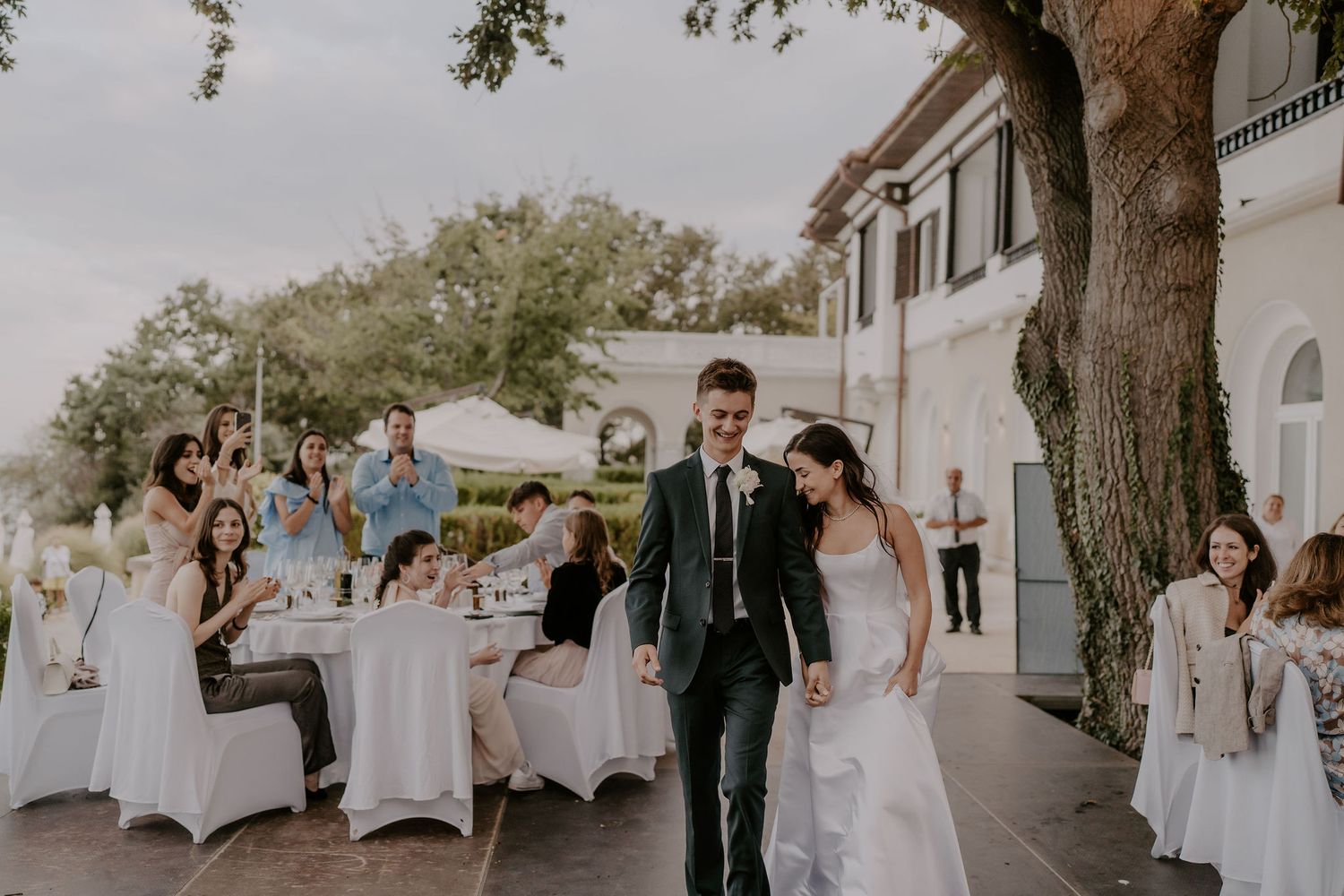 Wedding reception setup with white-covered chairs and tables arranged outdoors under trees.