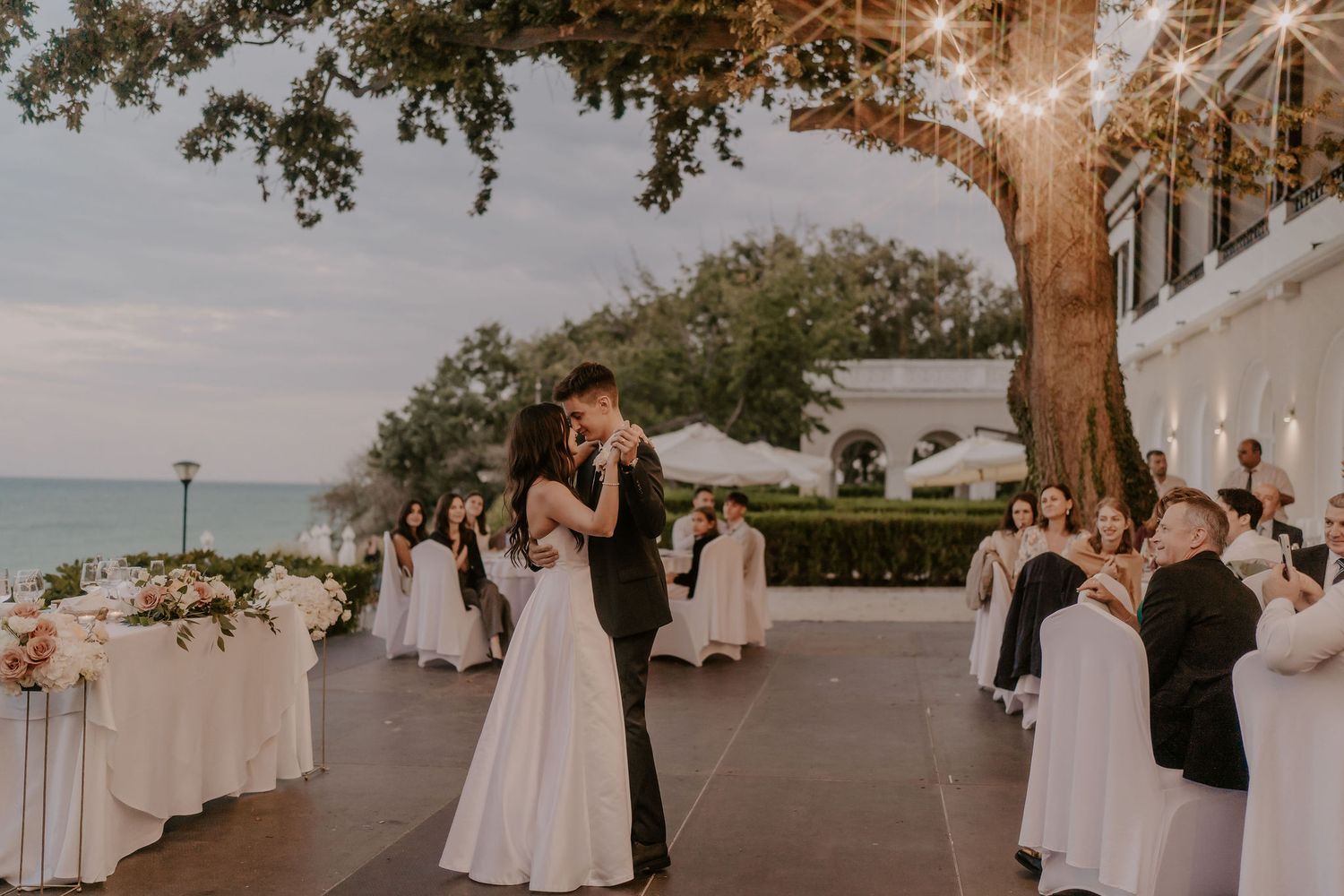 Newlyweds share their first dance at an elegant outdoor wedding reception by the ocean at sunset.