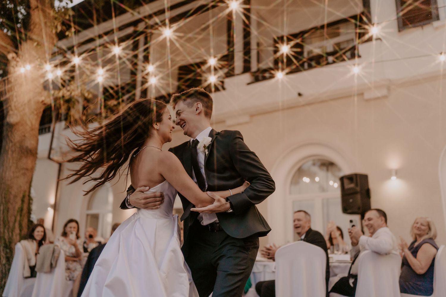 Couple dances energetically in an indoor venue with string lights and guests watching.