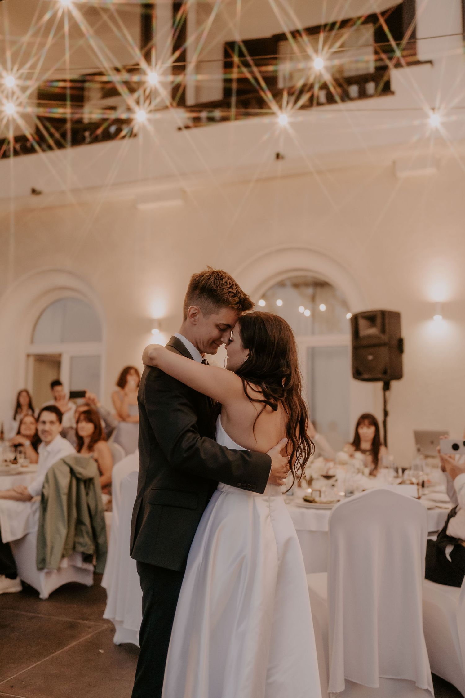 Couple embraces during their wedding dance under twinkling lights in an elegant indoor venue.