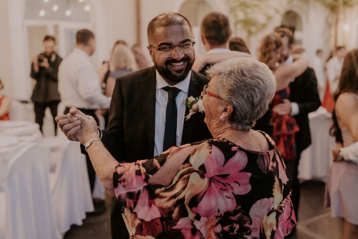 Wedding guest in a suit dances joyfully with woman in floral dress at reception.