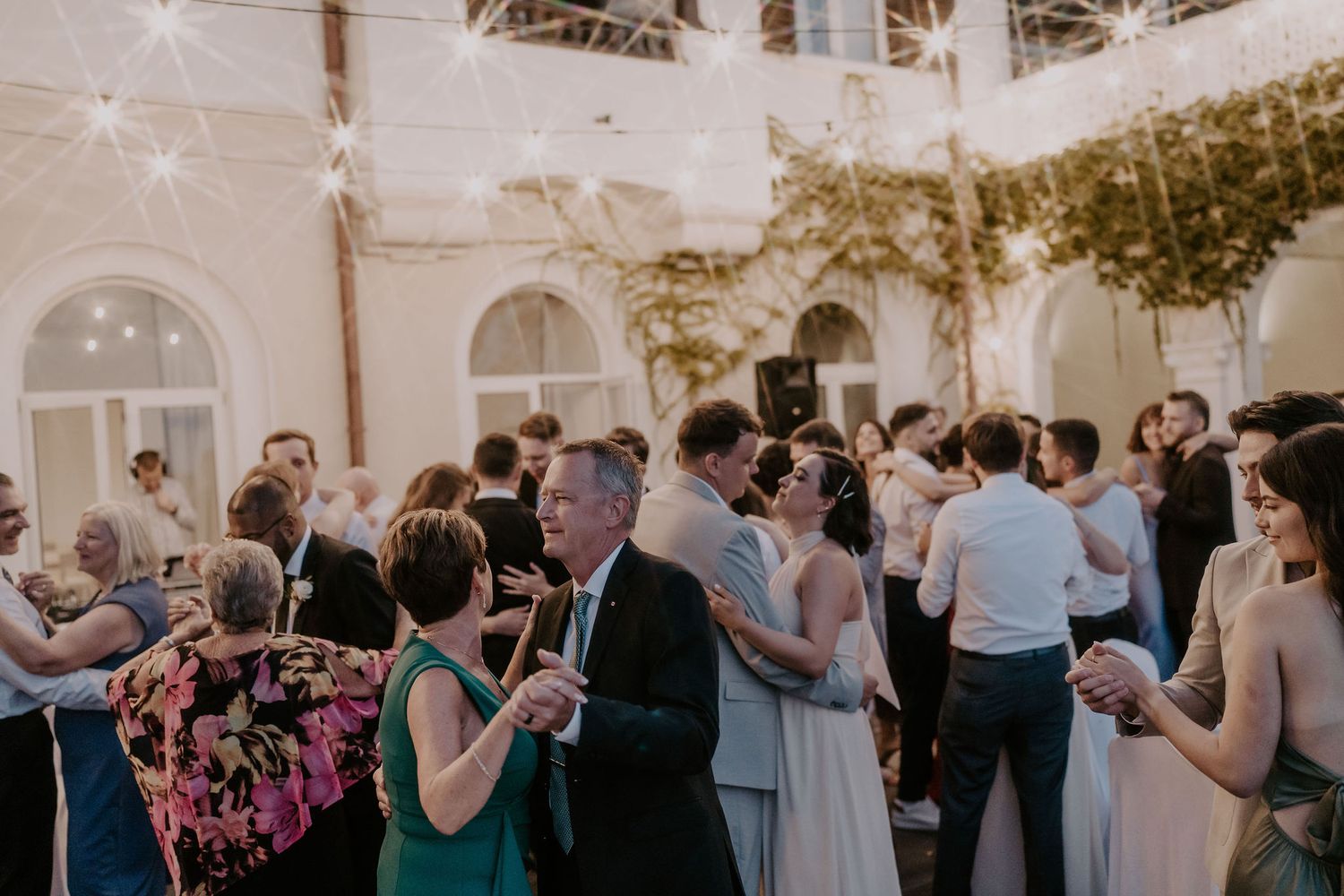 Wedding guests dancing and mingling in a beautiful courtyard with string lights and arched architecture.