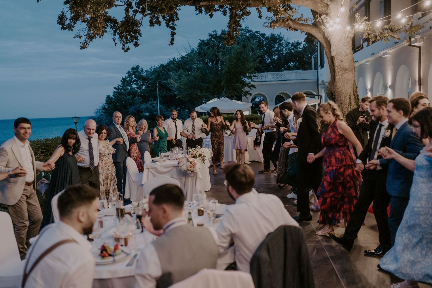 Wedding reception guests enjoy outdoor dining and dancing at dusk under trees.