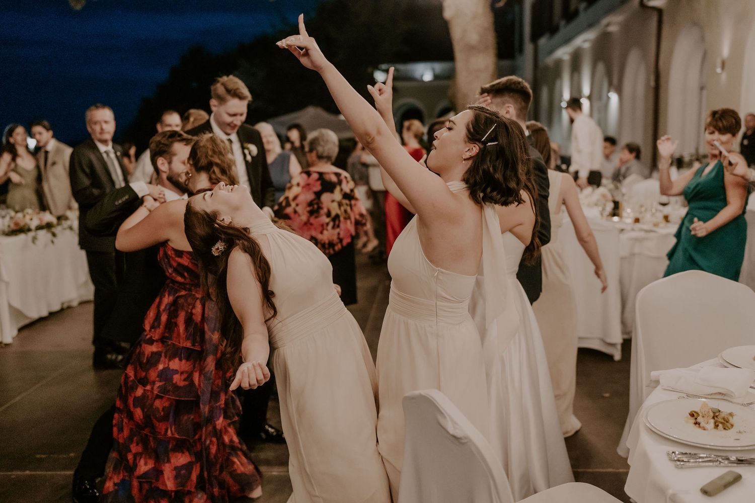 Wedding guests dance enthusiastically at an evening reception with decorative lighting.