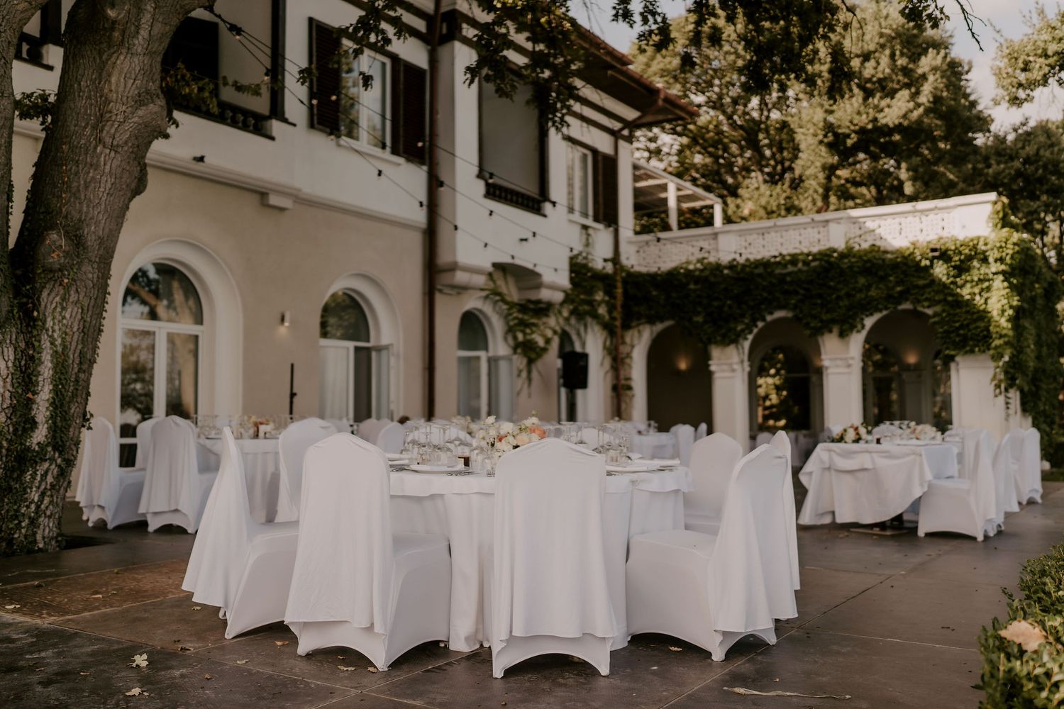 Elegant outdoor wedding reception setup with white-covered tables and chairs under a covered terrace.
