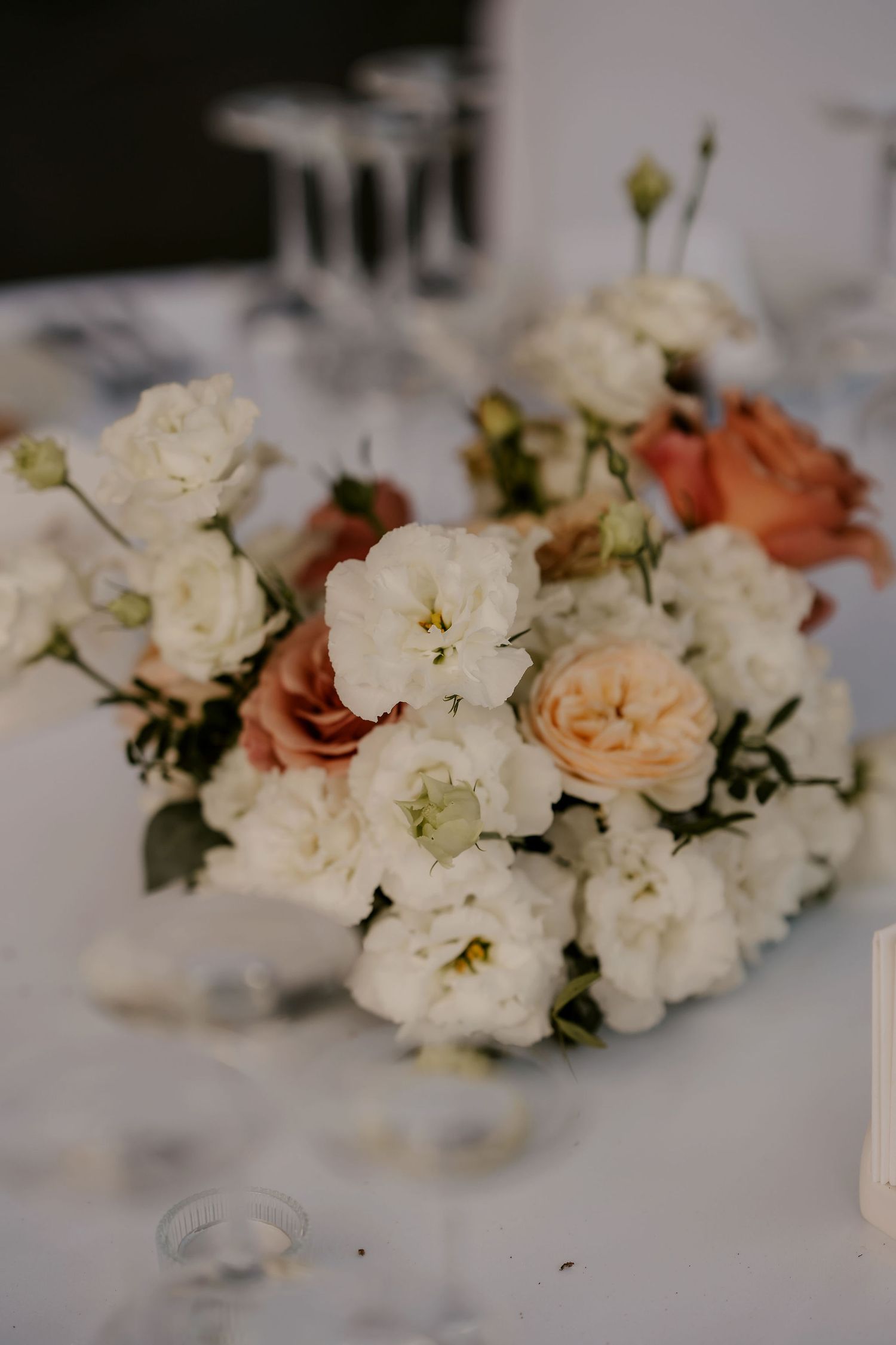 Delicate floral centerpiece featuring white and peach roses with greenery on a white tablecloth.