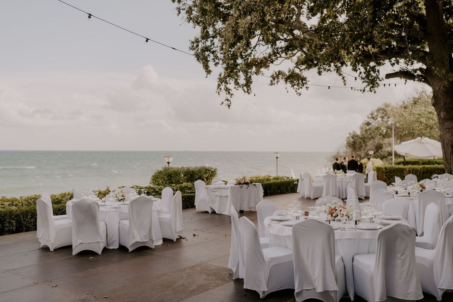Outdoor wedding reception area with string lights and white-covered tables near the ocean.