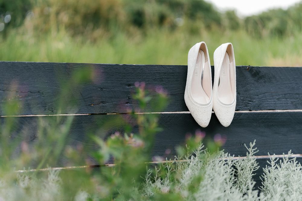 White bridal shoes displayed on a dark wooden bench outdoors with greenery in background.