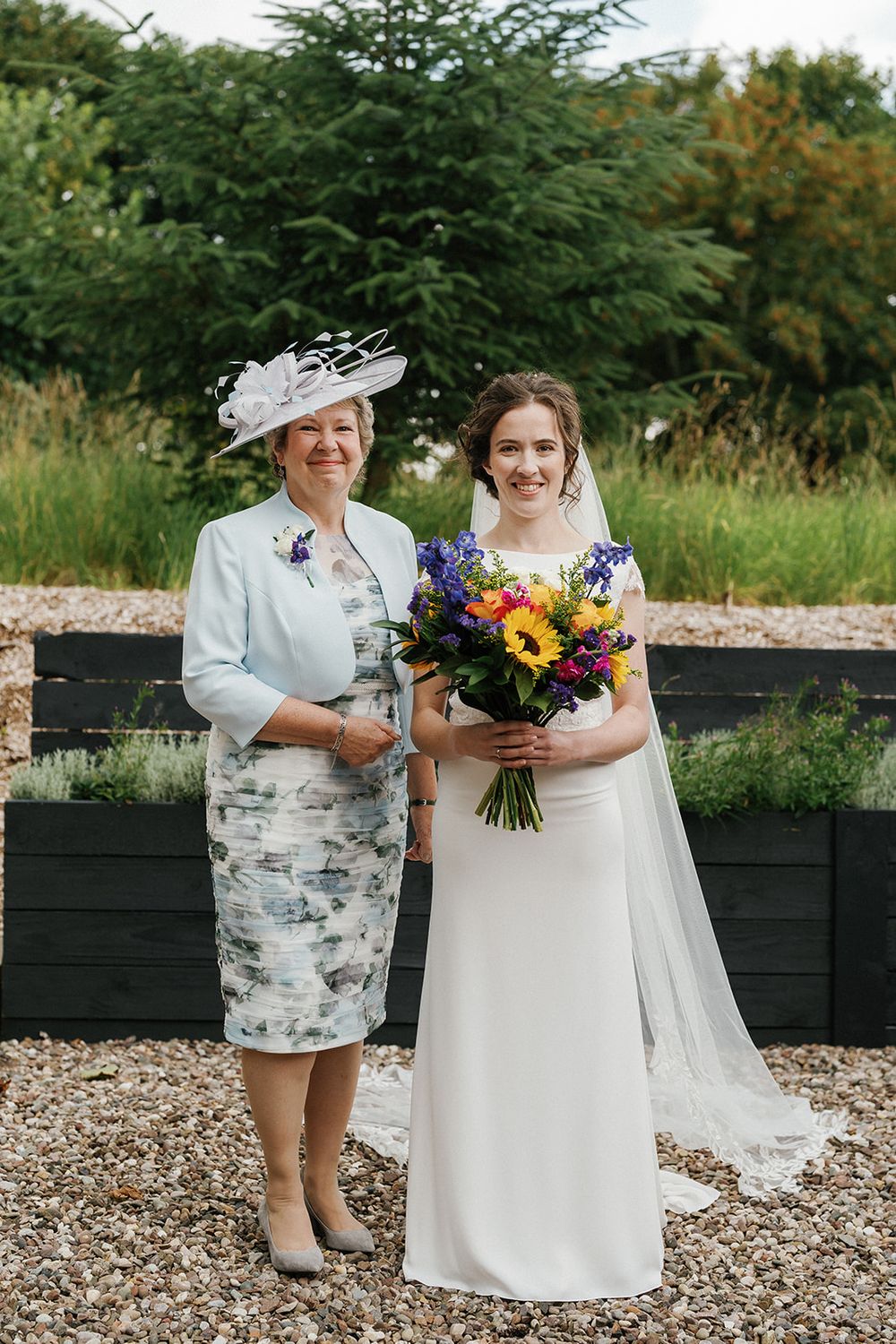 Wedding party members pose on gravel path with vibrant bouquets and traditional Scottish attire.