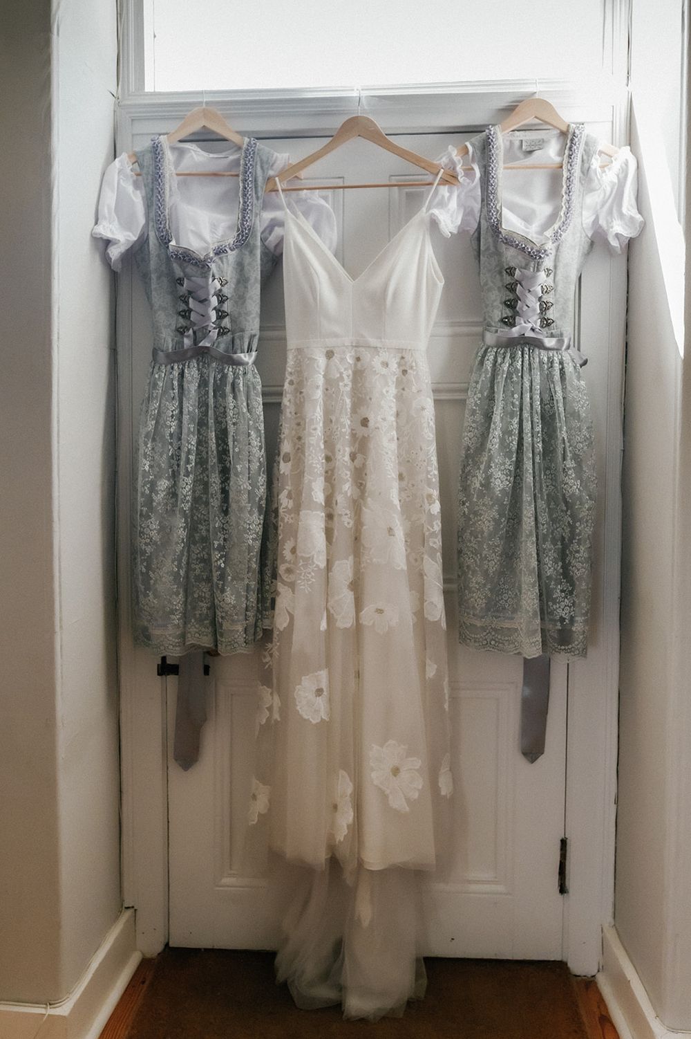 Traditional dirndl bridesmaid dresses in sage green hang beside an elegant white lace wedding gown on wooden hangers.