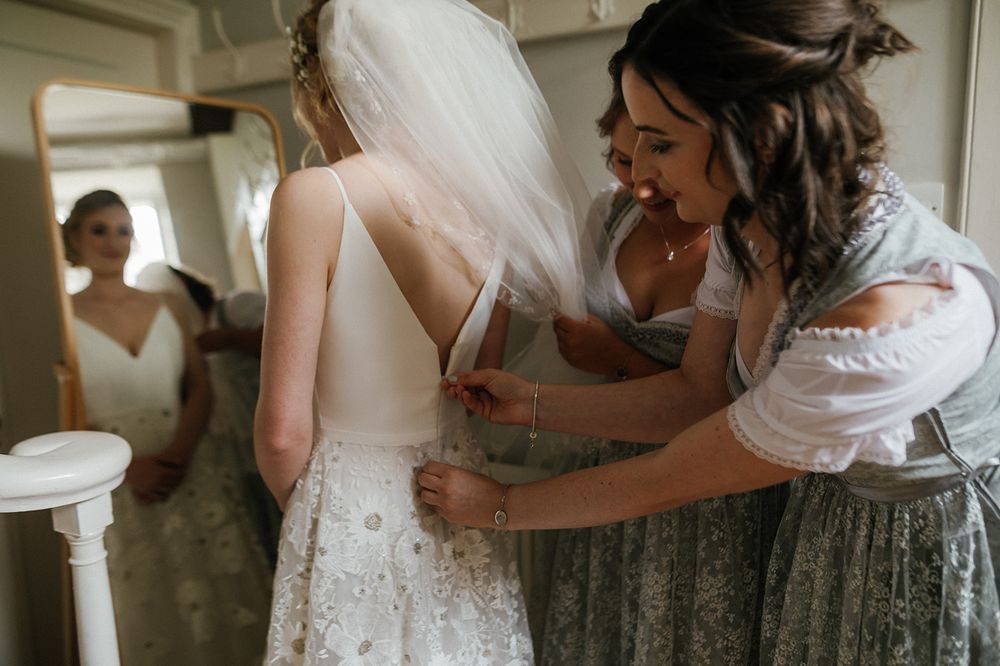 A wedding dress being fastened while bridesmaids in traditional dirndls assist with preparations.