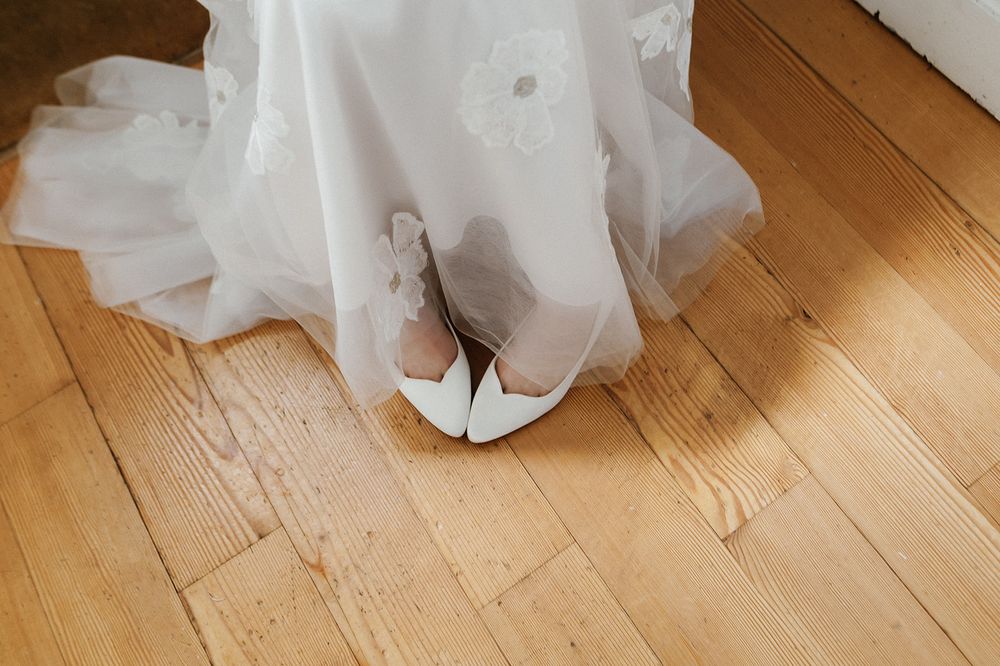 White bridal shoes peek out beneath a flowing wedding dress train on hardwood flooring.
