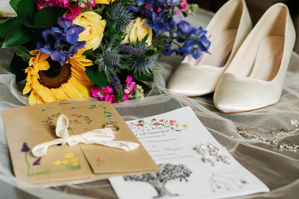 Wedding details including bridal shoes, colorful bouquet, and invitation cards arranged on a light surface.