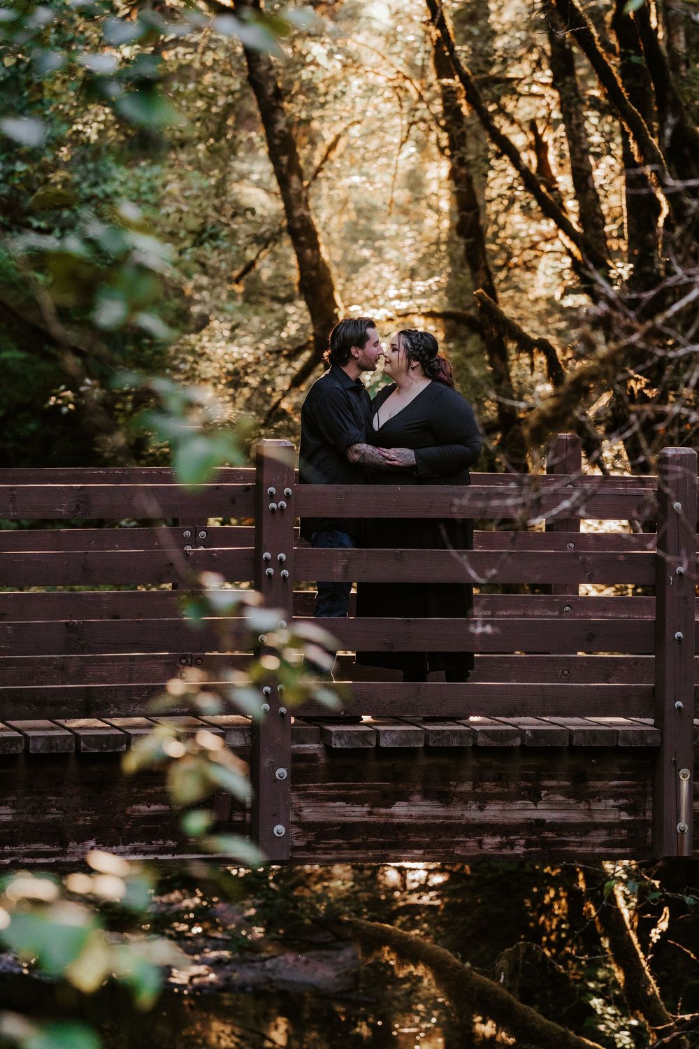 Couple shares a romantic almost-kiss on a wooden bridge surrounded by lush forest greenery.