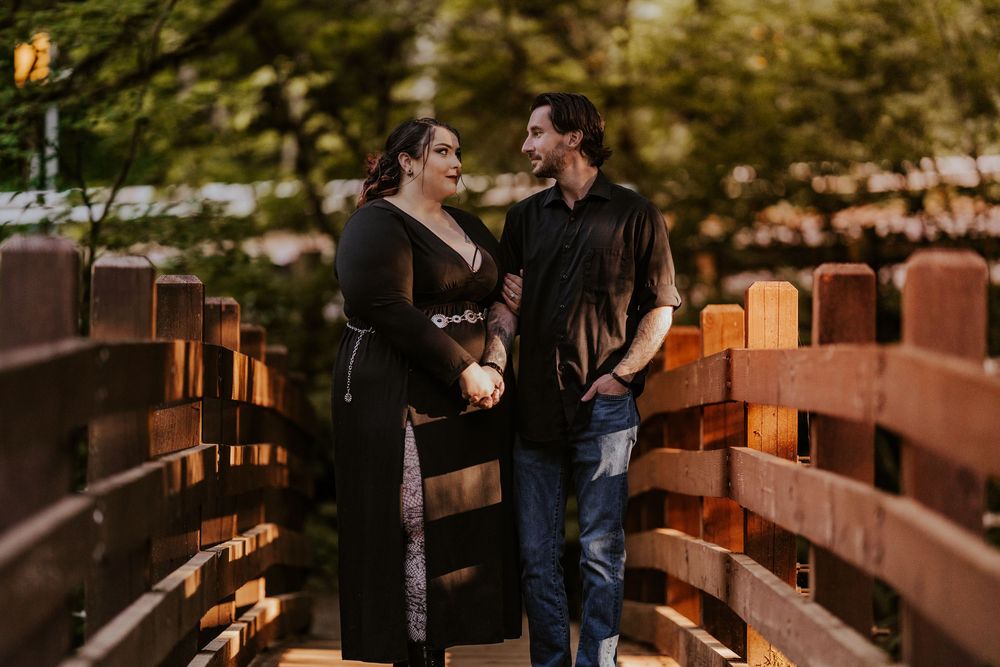 Couple in black outfits holding hands on a wooden bridge surrounded by lush green trees.