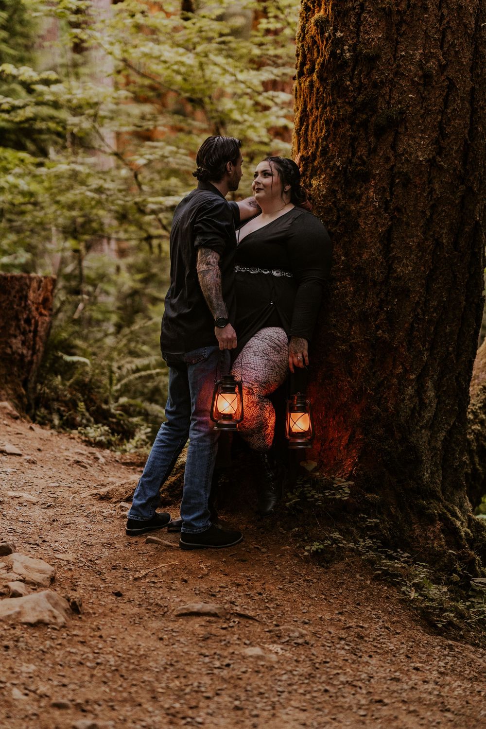 Romantic couple with glowing lanterns leaning against a large tree in a forest setting.