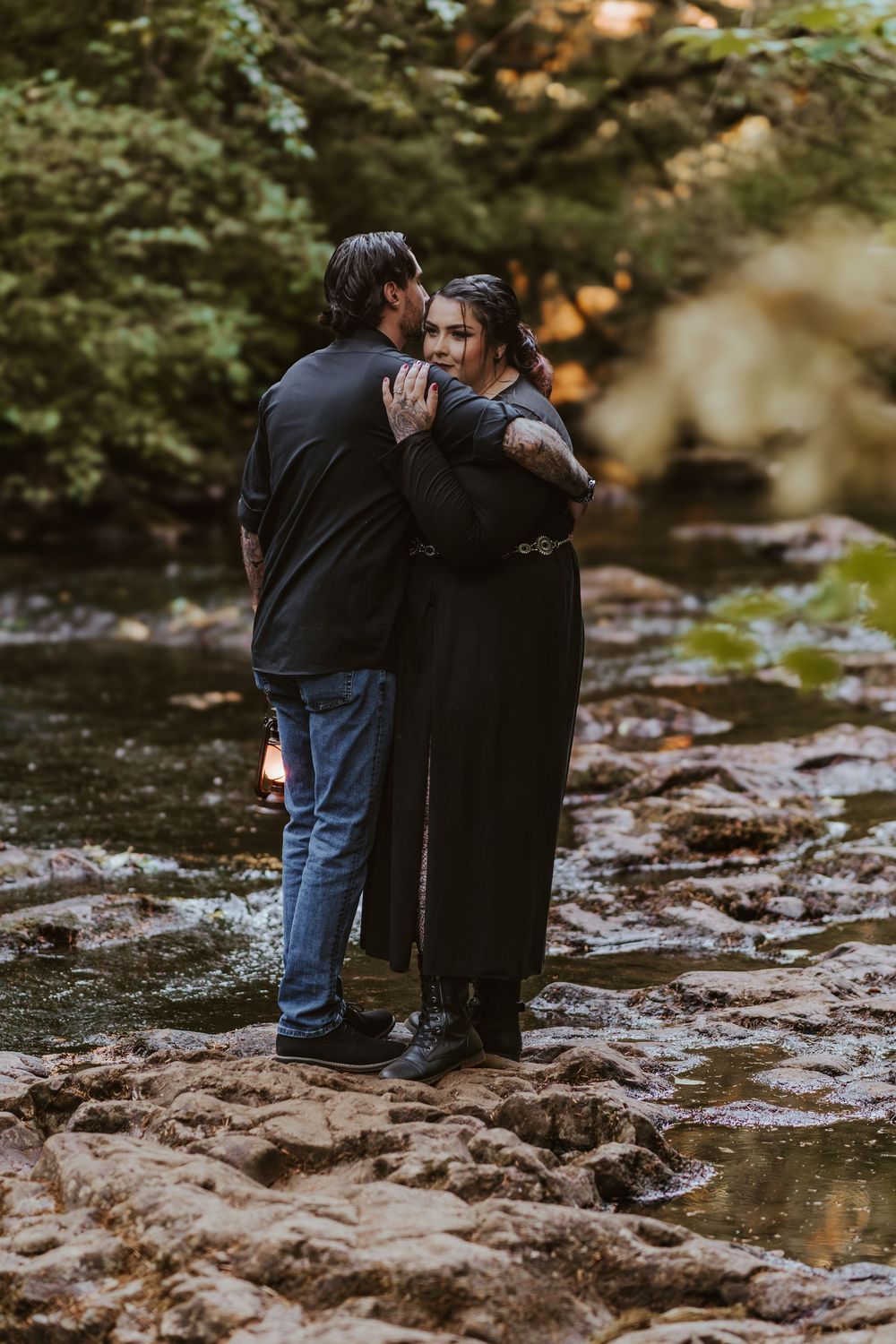 Couple embracing on rocky creek bed surrounded by green forest trees.