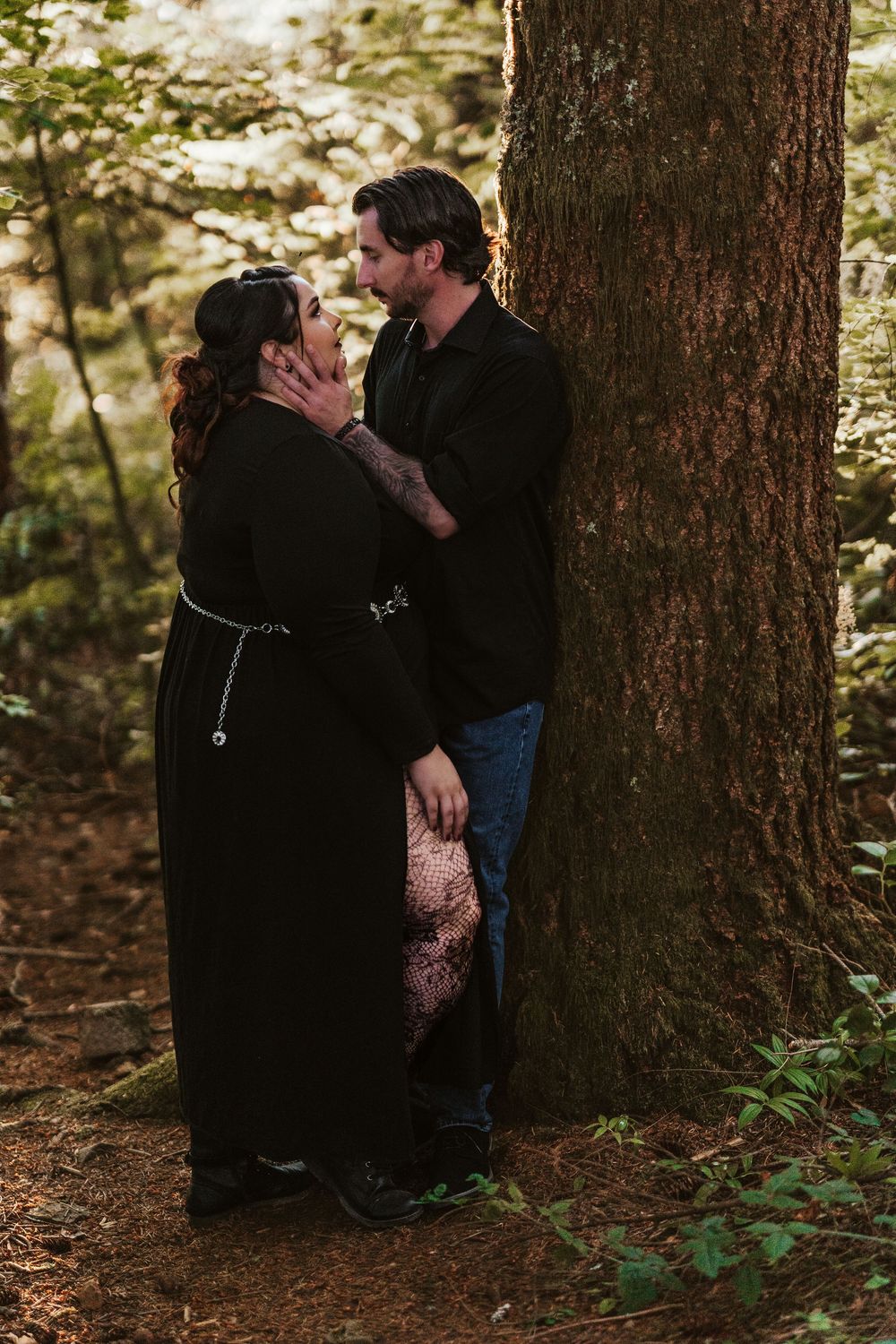 Couple in black outfits sharing an intimate moment leaning against a large forest tree.