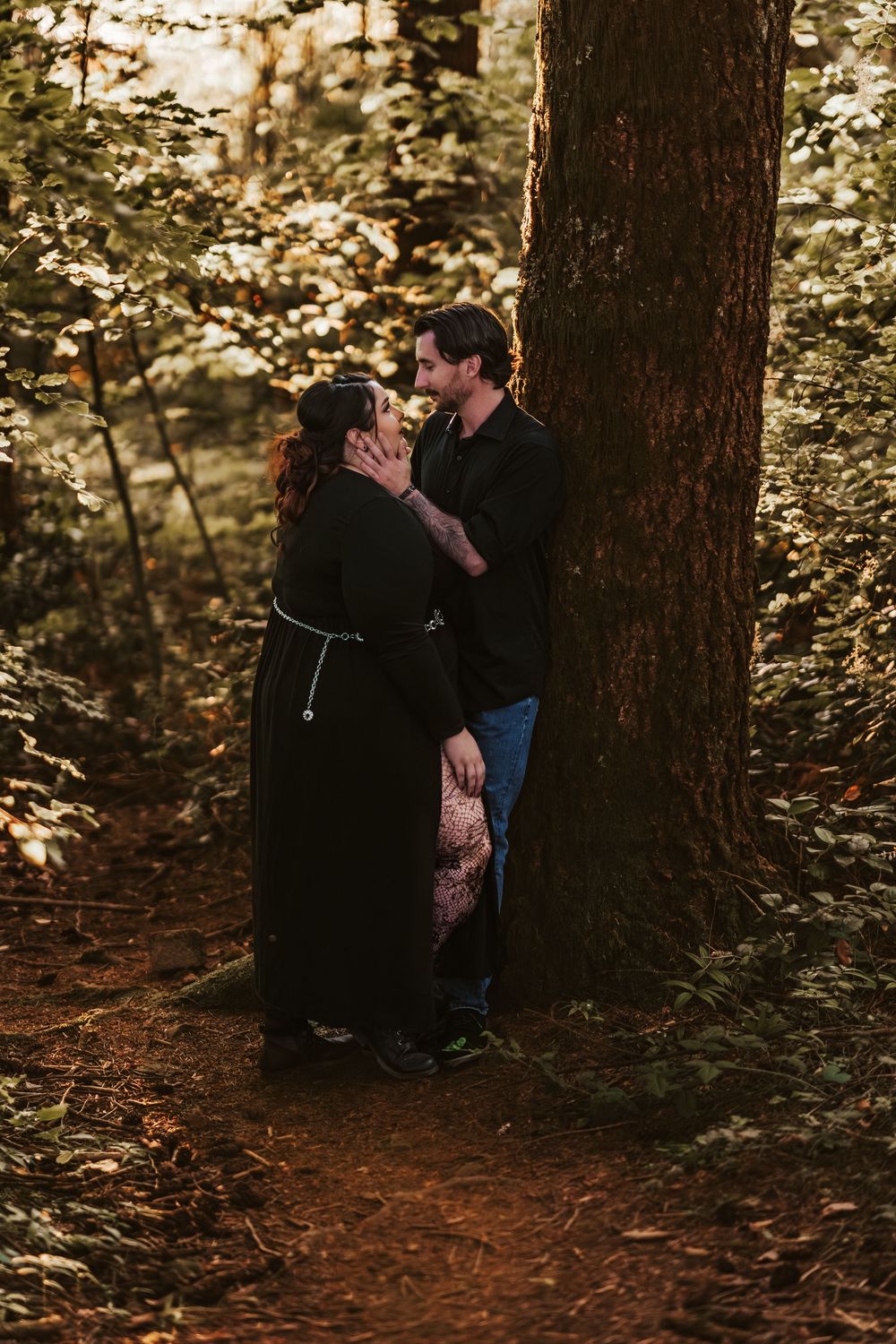 Couple nearly kissing while leaning against a tree in golden sunlit forest.