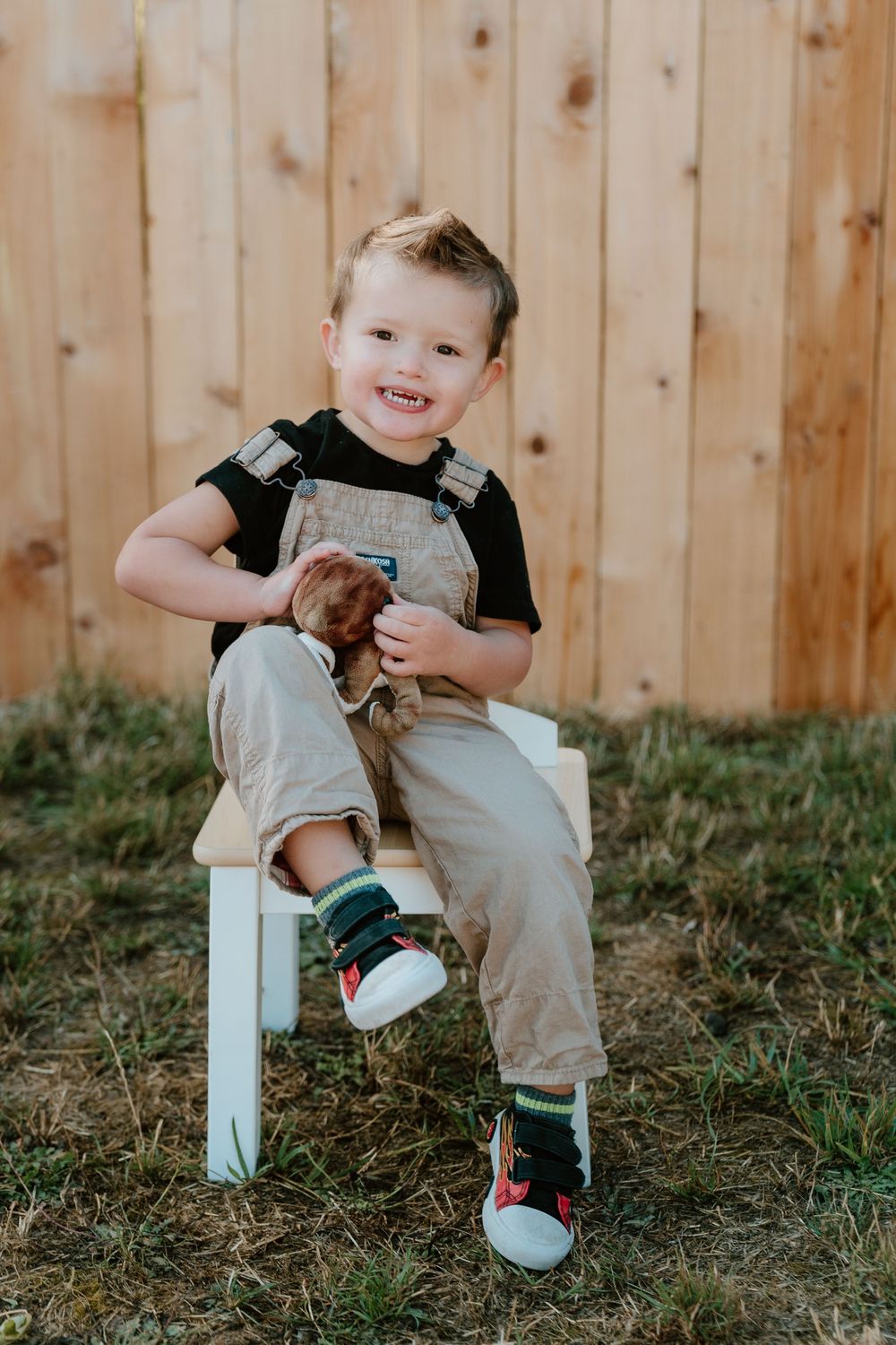 A cheerful toddler boy in tan overalls sits on a small white bench holding a stuffed animal by a wooden fence.