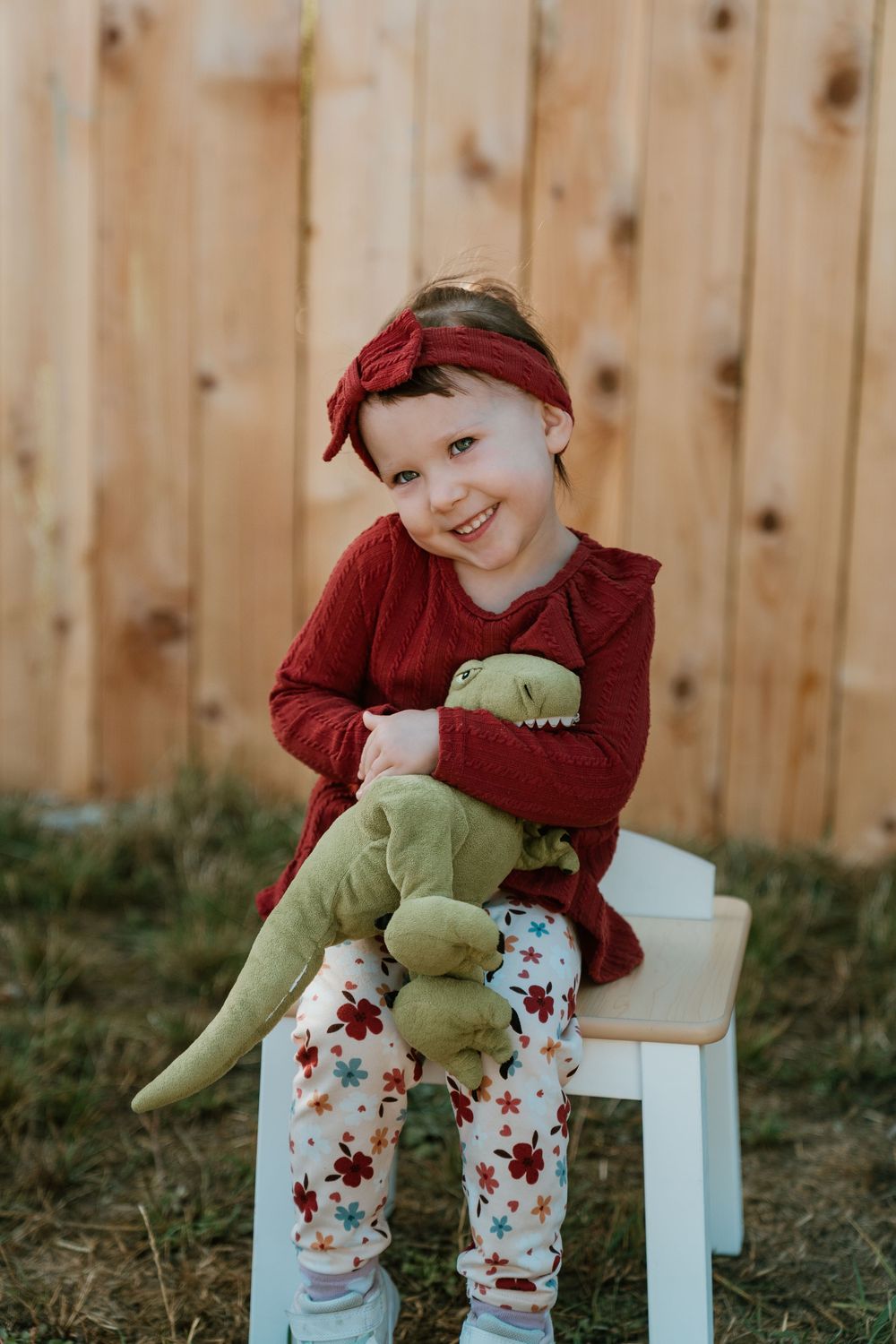 A smiling little girl in a red outfit and headband sits on a white bench hugging a green dinosaur stuffed toy.