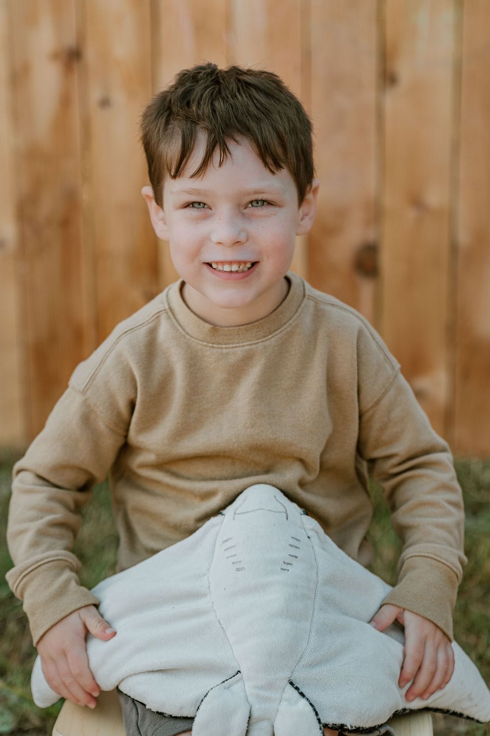 A happy young boy in a tan sweatshirt sits on a wooden bench holding a gray stuffed elephant toy.