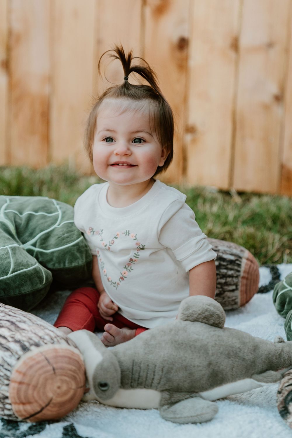Smiling baby girl with dark hair sitting on blanket outdoors with stuffed animal toys and woodland-themed pillows.
