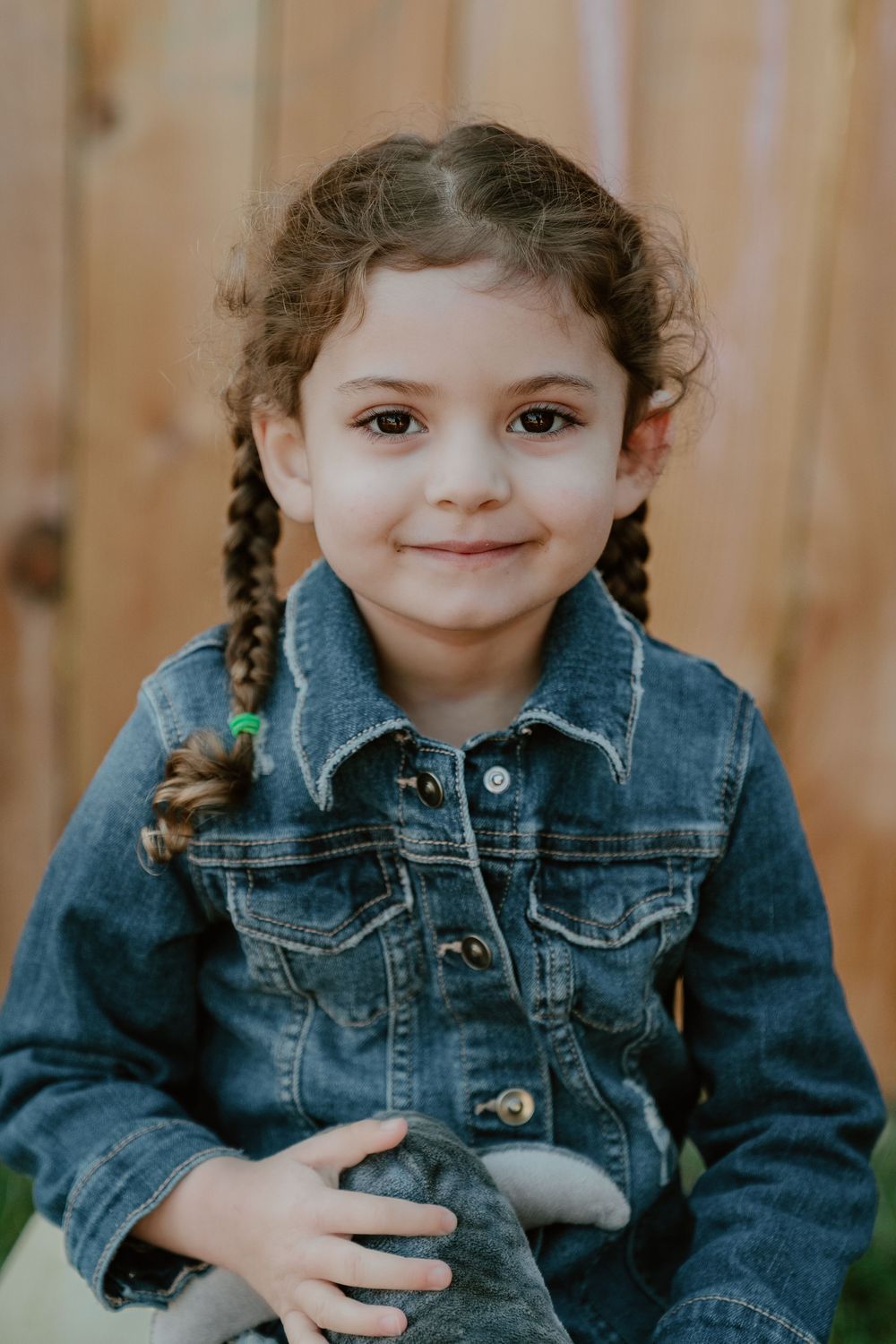 Adorable toddler girl with braided pigtails wearing denim jacket holding stuffed animal toy outdoors.