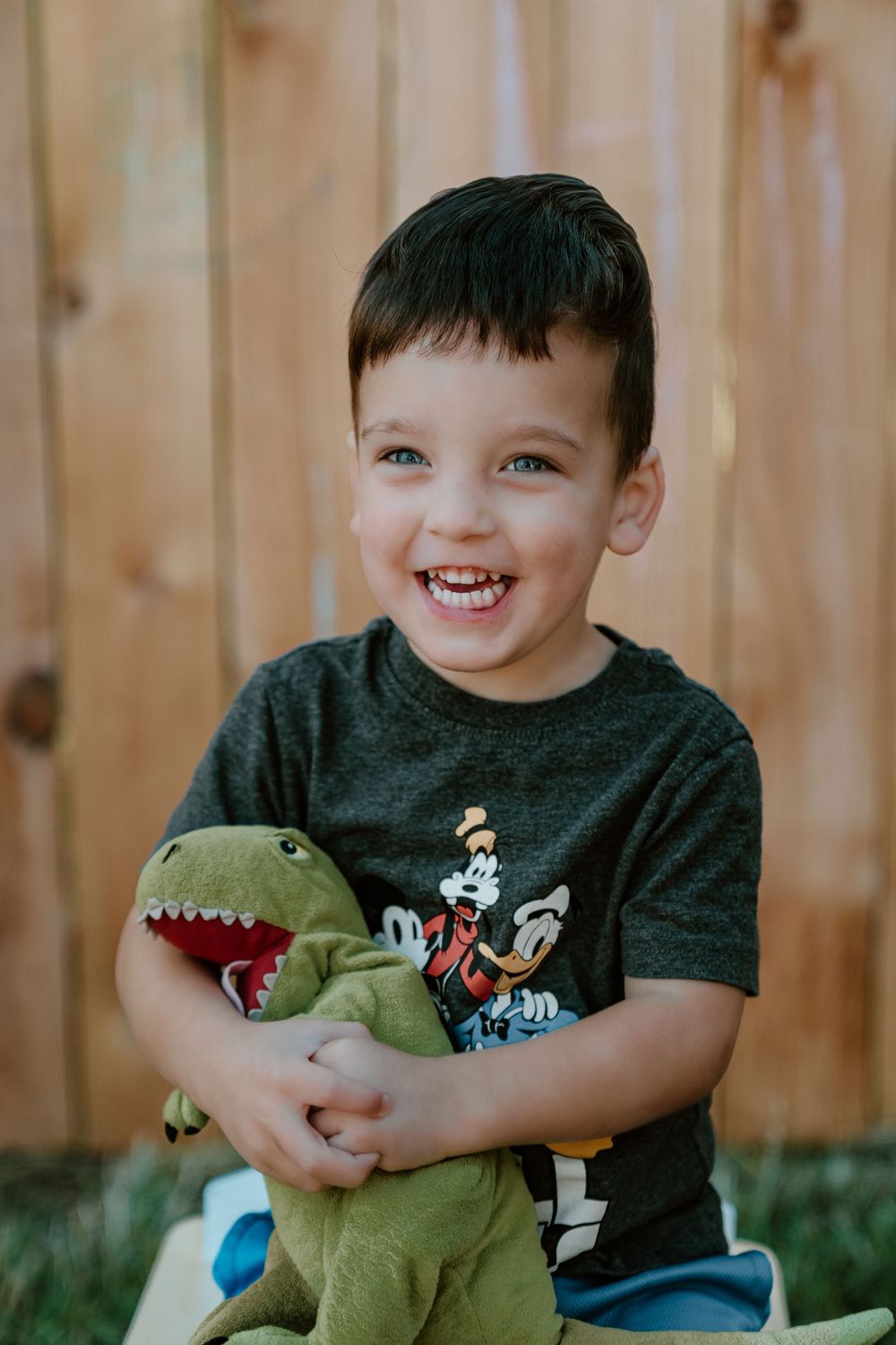Happy young boy with blue eyes laughing while holding green dinosaur stuffed animal toy outdoors.