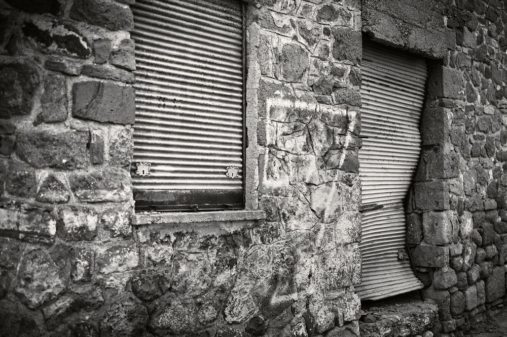 Two weathered windows with metal shutters set in an old stone wall captured in black and white.