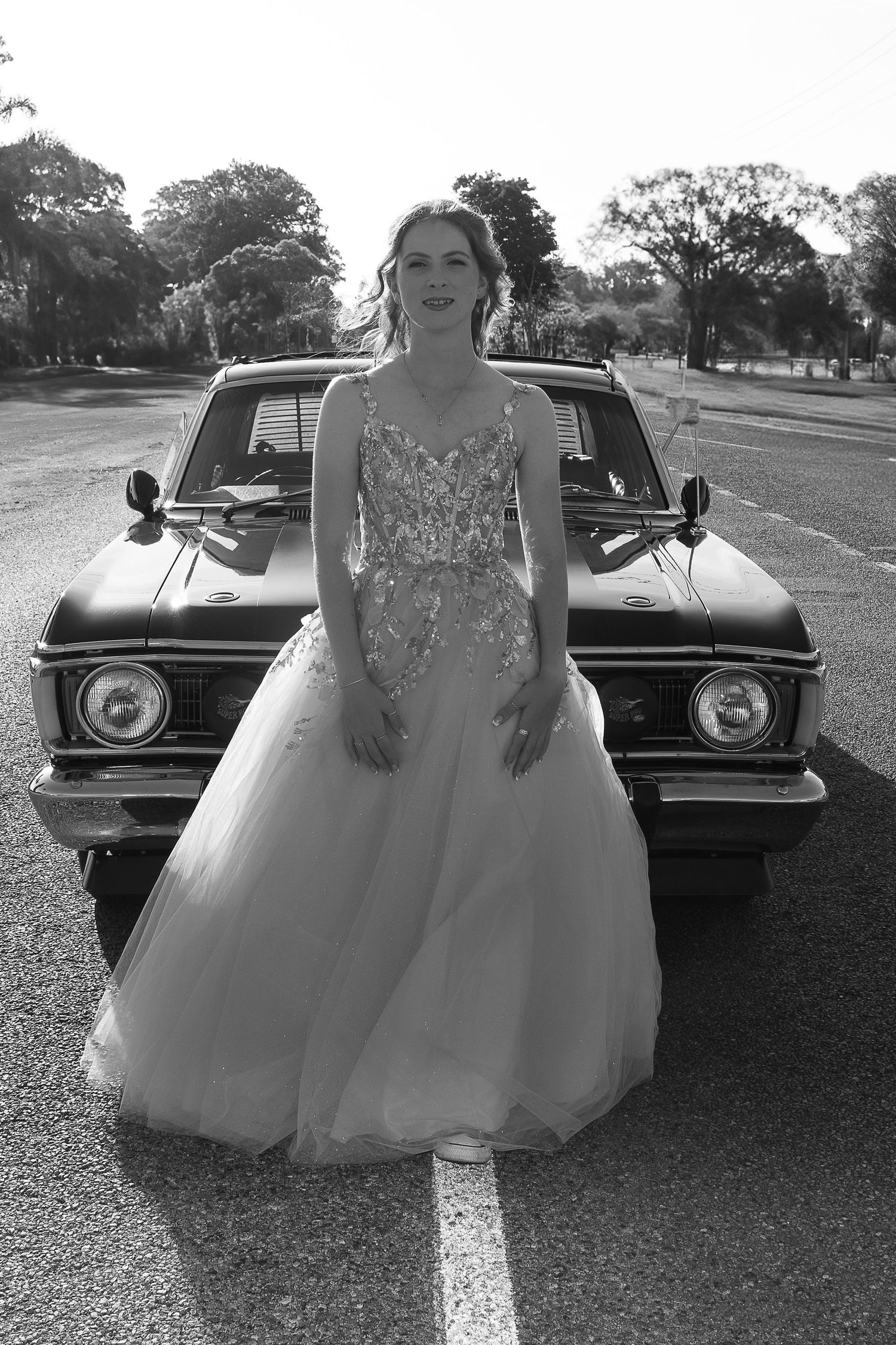 A person in a sparkly white ball gown stands in front of a vintage black car on a sunny day.