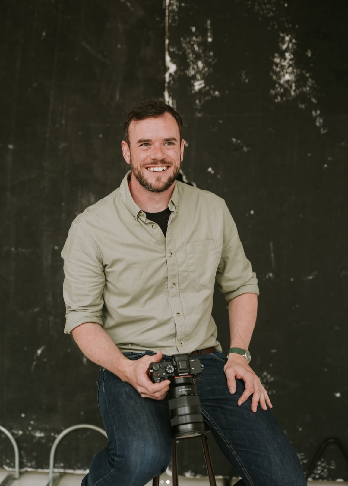 Professional photographer smiling while posing with camera equipment against dark textured wall.