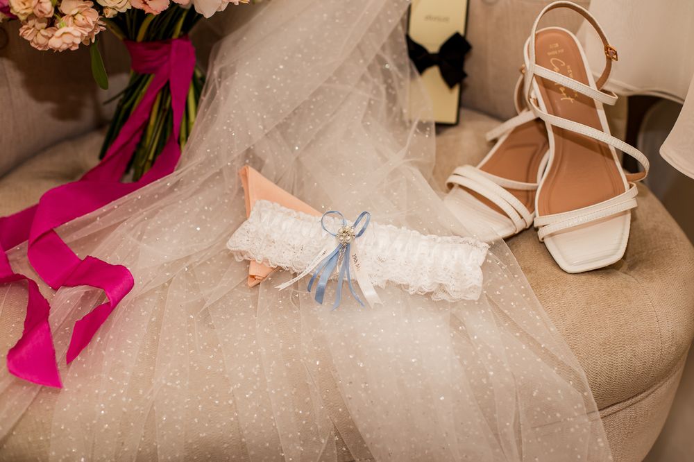 Wedding accessories including bridal shoes, ribbons and floral arrangements laid out on beige fabric.