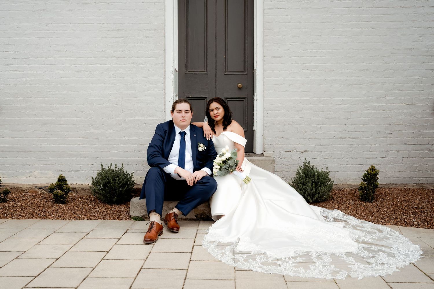 A bride in a white lace gown with long train sits beside her companion in a navy suit on stone steps.