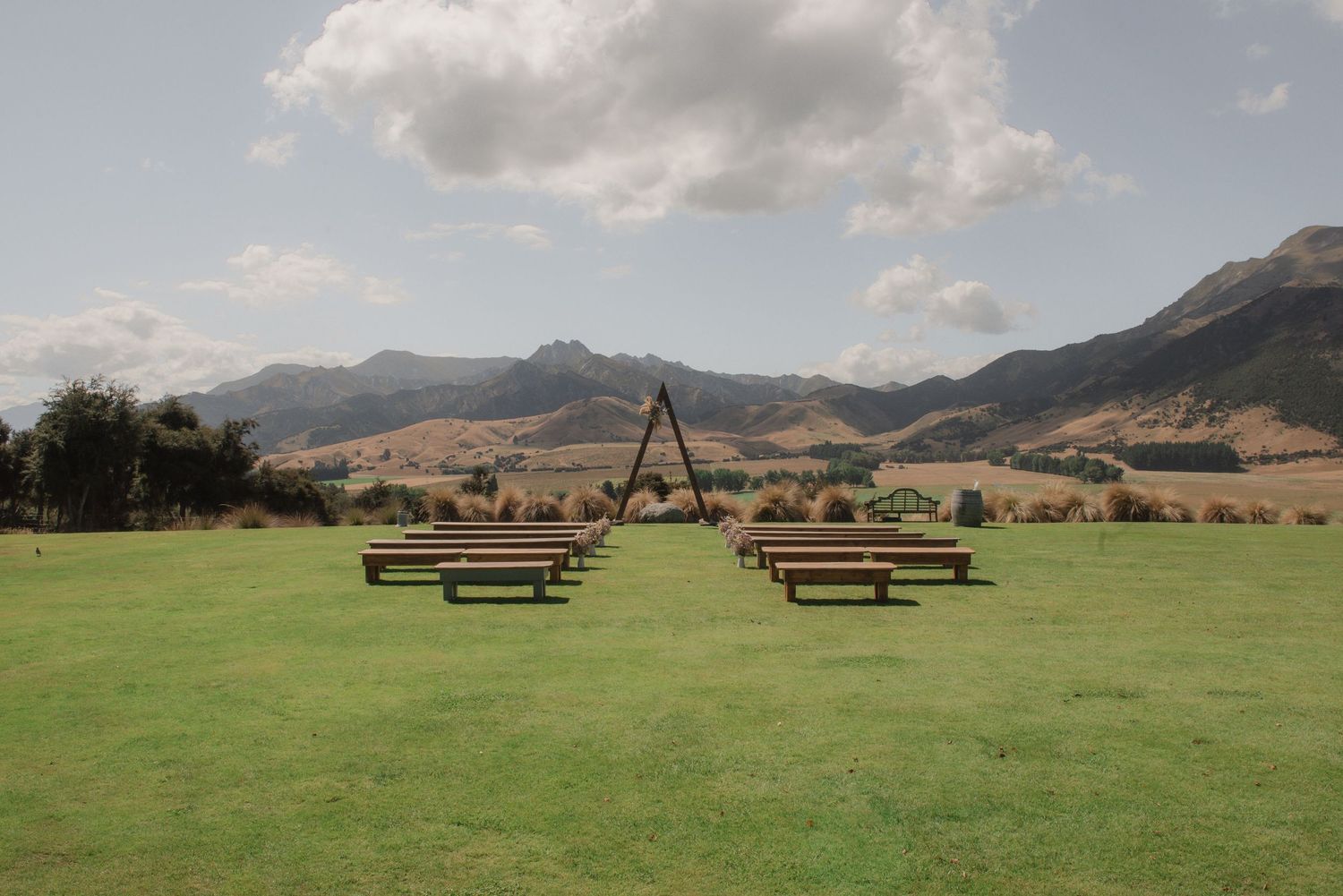 A panoramic view of rolling green hills with wooden benches overlooking a scenic mountain landscape on a sunny day.