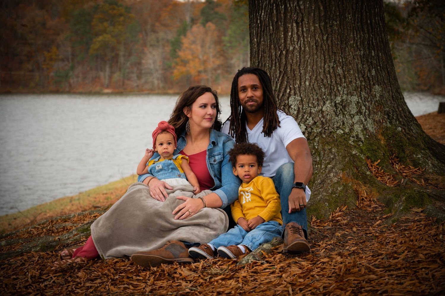 Family sitting together under large tree by lake during autumn with colorful fall foliage in background.