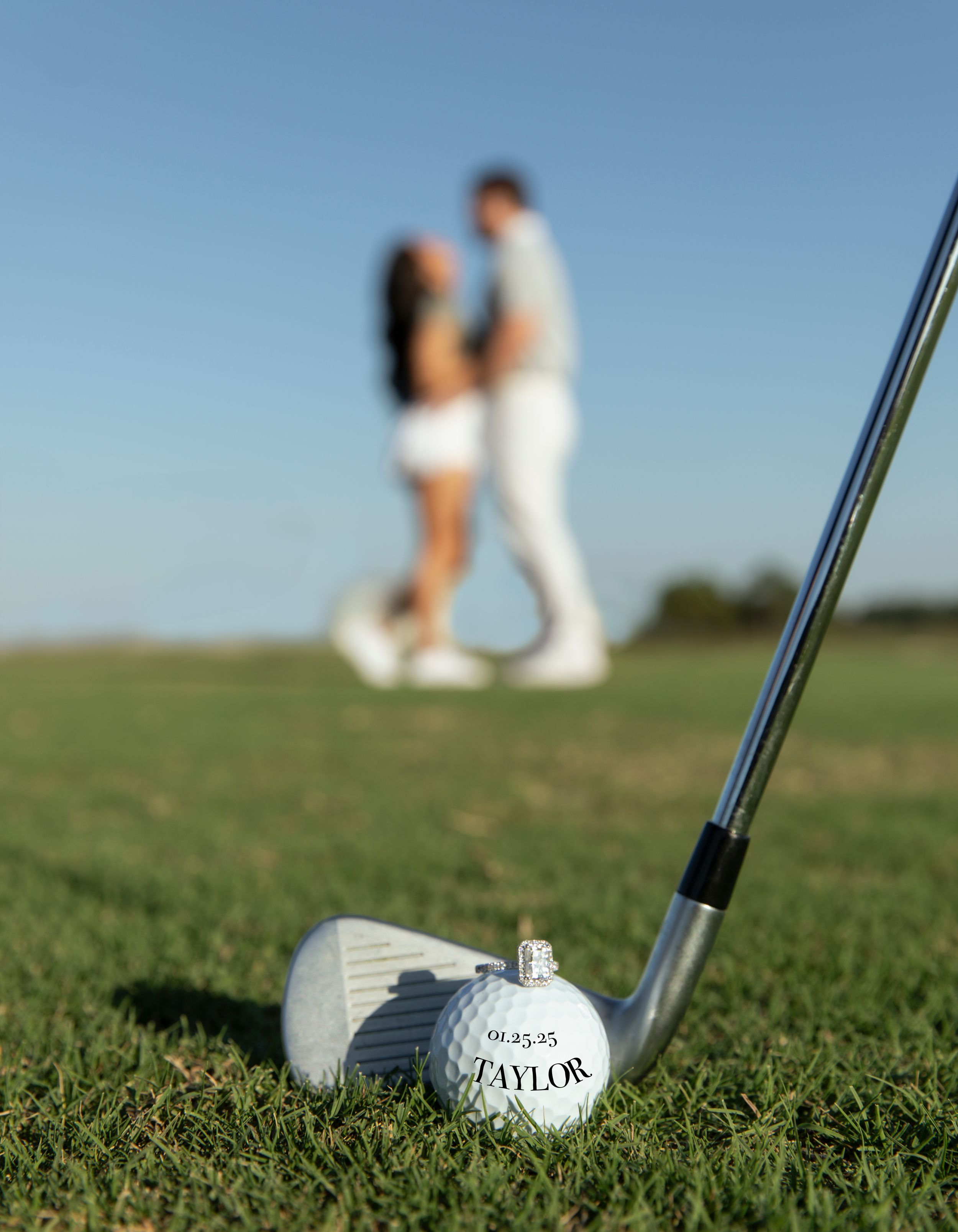 A golf club and ball on grass with a romantic couple embracing in the blurred background at a golf course.