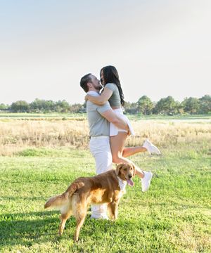 A couple embracing in a sunny meadow with their brown dog running playfully in the foreground.