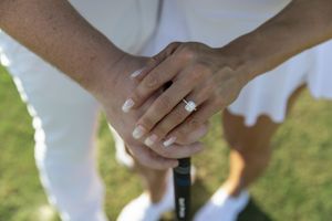 Hands clasped together wearing a diamond engagement ring with green grass in the soft-focus background.