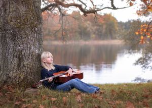 En flicka med sin gitarr sitter under ett träd, med en sjö i bakgrunden.