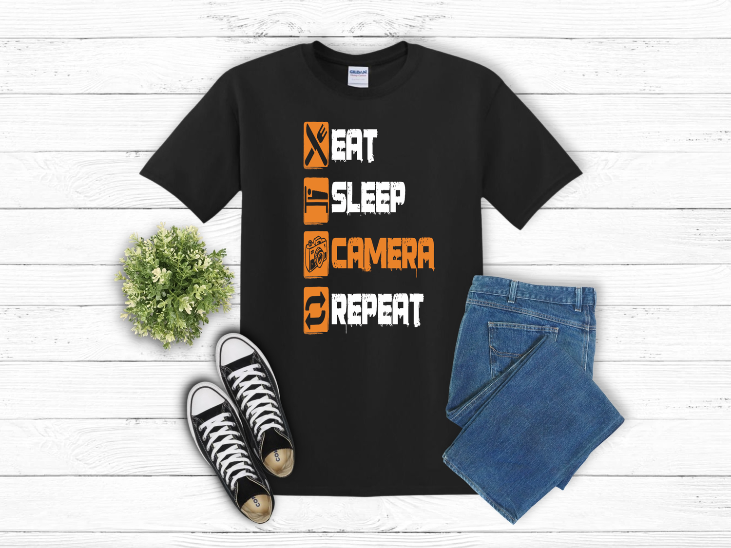 Black t-shirt with orange text 'Eat Sleep Camera Repeat' displayed with jeans and black sneakers on white wooden surface.