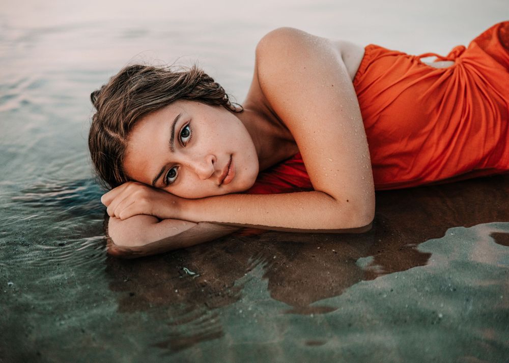 A figure in a vibrant orange dress lies serenely at the water's edge during sunset, creating reflections on the wet sand.
