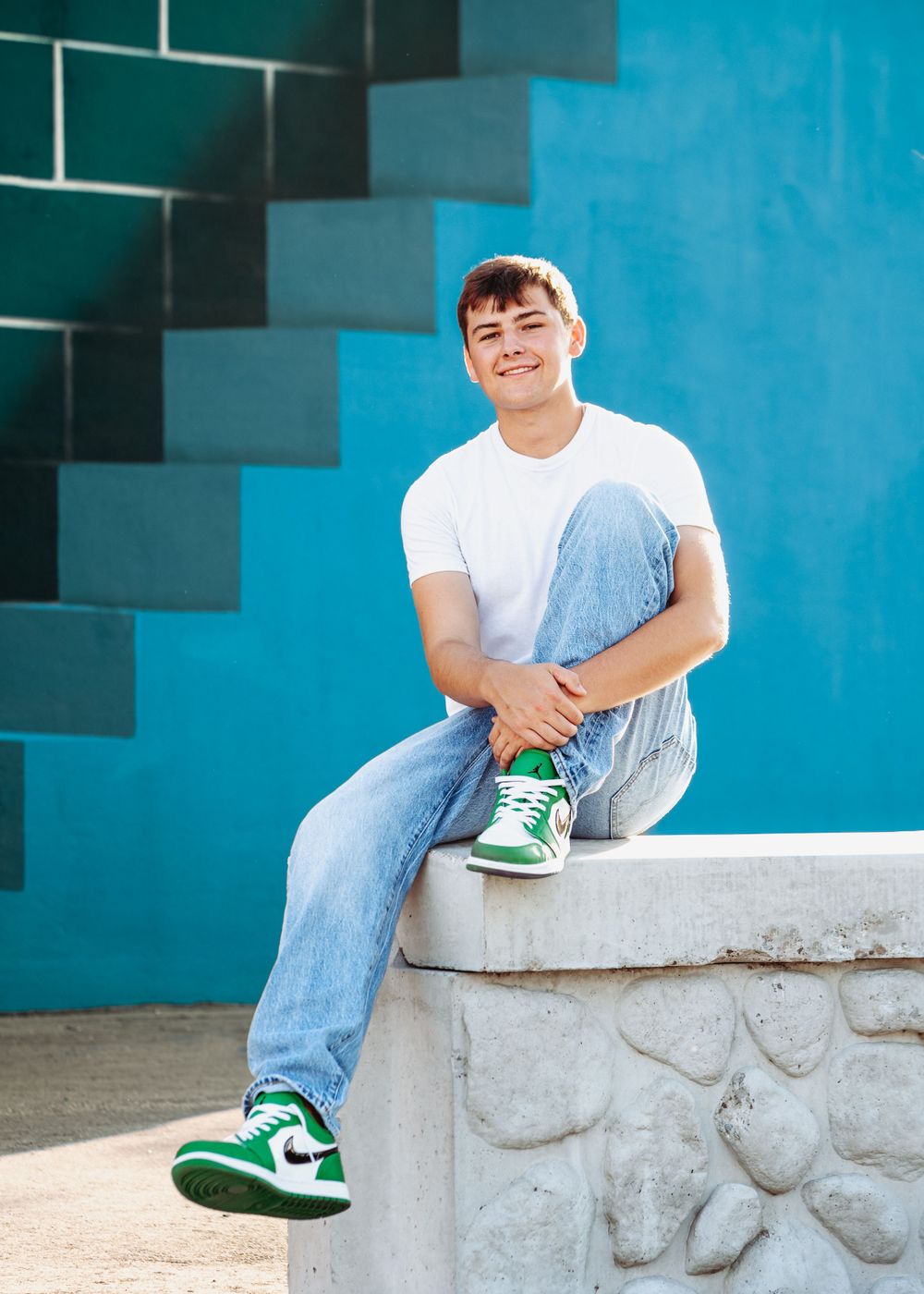 Someone in light denim jeans and white t-shirt sits casually on concrete ledge against turquoise geometric wall backdrop.