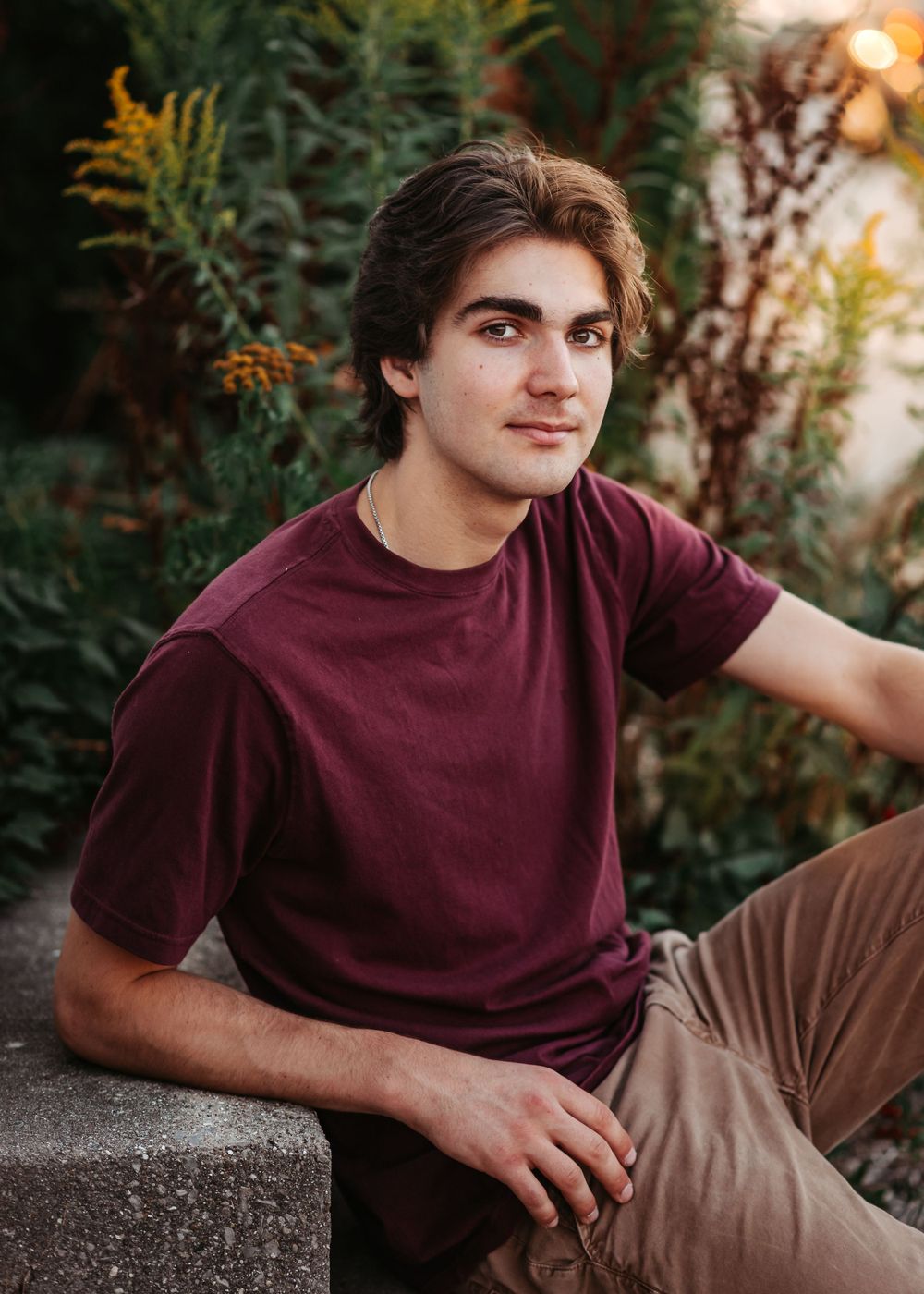 A young person in a burgundy t-shirt and khaki pants relaxes outdoors at sunset with greenery in the background.