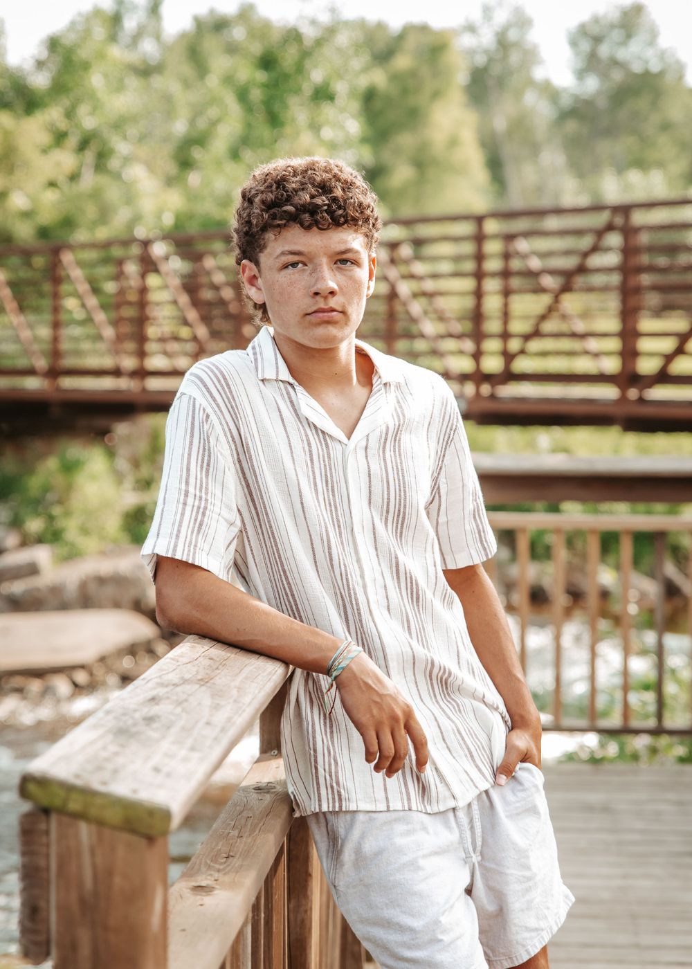 A person in a white striped button-down shirt poses against a rustic wooden bridge backdrop on a sunny day.